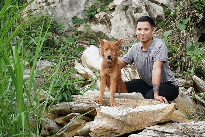 Người ngồi cạnh một chú chó màu nâu trên các tảng đá, xung quanh là cây cối xanh tươi.
