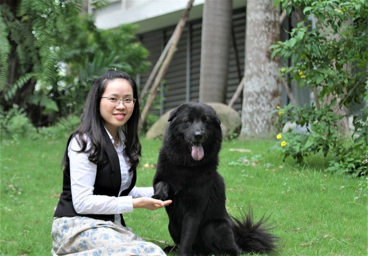 Người phụ nữ ngồi cạnh chó đen lớn trong vườn, cả hai đều nhìn vào máy ảnh, xung quanh là cây cối xanh tươi.